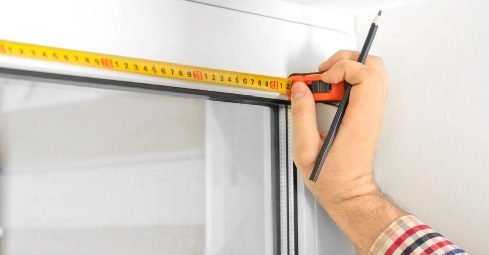 A person's wrist and hand as they press yellow measuring tape up to a window and hold a black pencil.