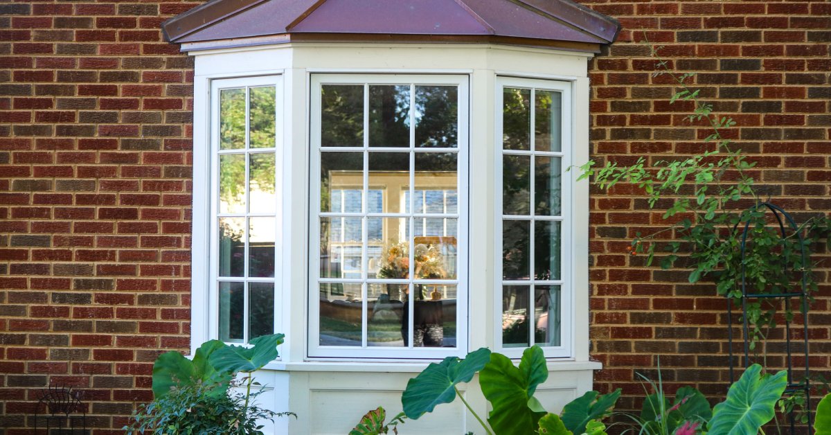A white bay window installed to the surface of a brick house reflects the view of trees and flowers.