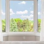 A window in a room adorned with white and gray curtains displays a view of cloudy blue skies and trees.