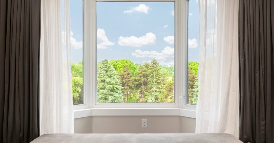 A window in a room adorned with white and gray curtains displays a view of cloudy blue skies and trees.