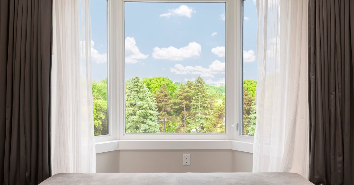 A window in a room adorned with white and gray curtains displays a view of cloudy blue skies and trees.