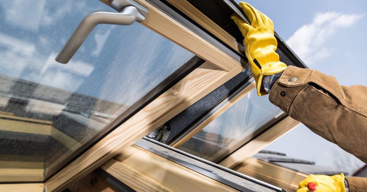 A person wearing yellow work gloves holds open a pair of windows that have silver handles and wooden frames.