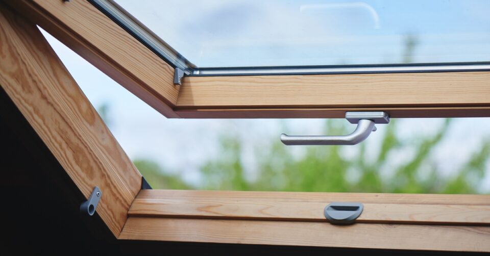 An open window with a silver handle and wooden frame. A blue sky and green plant leaves are visible outside.