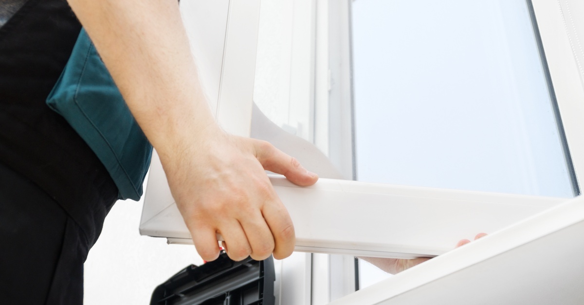 A person's hands holding an opened window with a white frame, installed against a plain white surface.