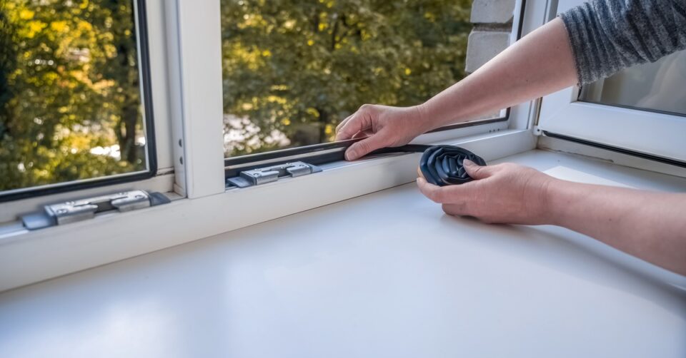 A person uses both hands to apply dark rubber sealing tape to the surface of an interior window frame.