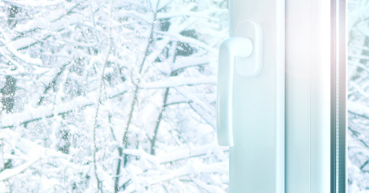 Tree branches covered in snow are visible through the glass of a white window with a handle attached to it.