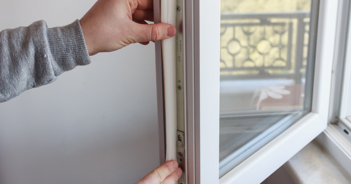 A person wearing a gray shirt uses both hands to apply insulating tape to an open window with a white frame.