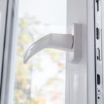 A close-up of an open white vinyl window with a handle on one side. Trees and sunlight are visible through the glass.