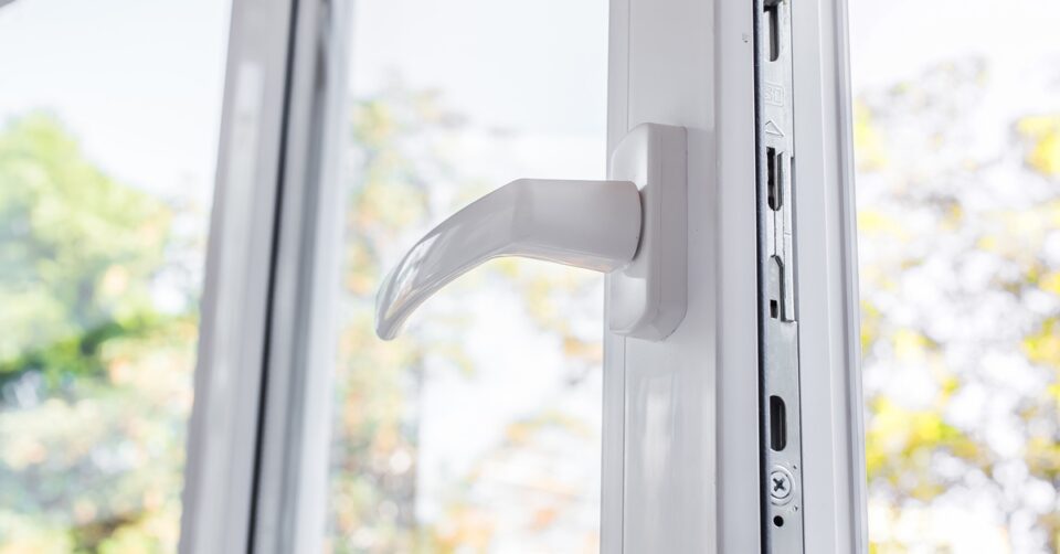 A close-up of an open white vinyl window with a handle on one side. Trees and sunlight are visible through the glass.