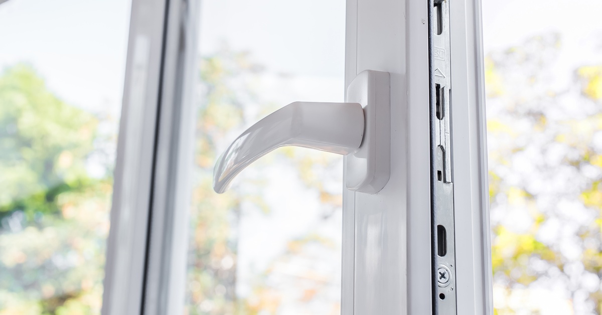 A close-up of an open white vinyl window with a handle on one side. Trees and sunlight are visible through the glass.