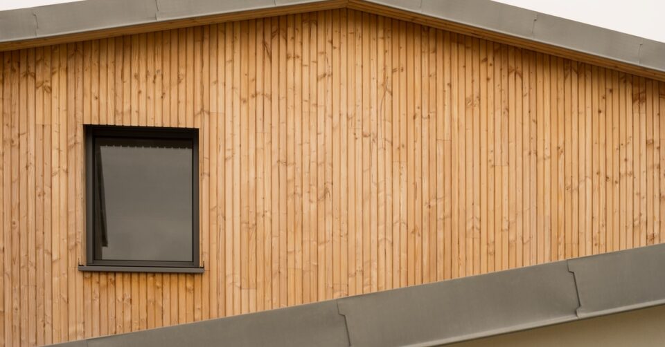 Part of a building with a brown metal roof, wooden siding, and a window with a black metal-clad frame.