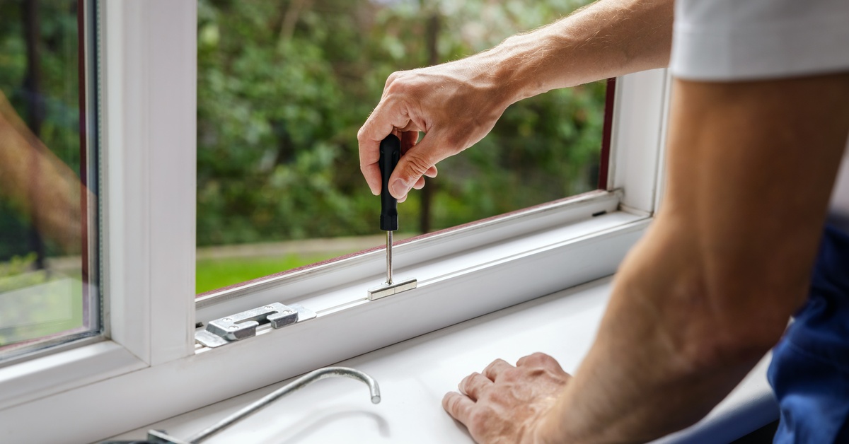 A person wearing white and blue clothes holds a tool in their right hand and presses it on the white frame of a window.