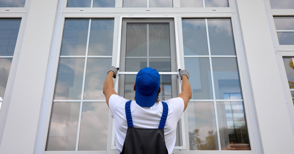 The back of a person wearing a blue jumpsuit and hat installing a window screen on the outside of a home.