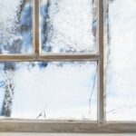 A close up of a frosty window with a wooden frame. Ice crystals have formed on the window panes, and there's snow outside.