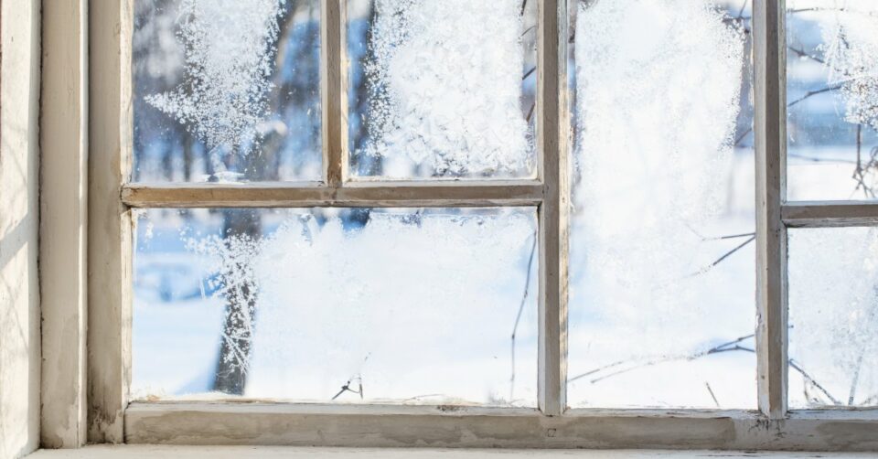 A close up of a frosty window with a wooden frame. Ice crystals have formed on the window panes, and there's snow outside.