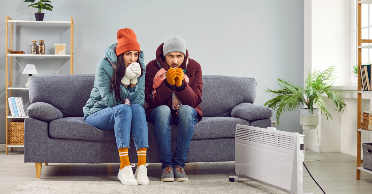 Two people sit on a couch wearing winter jackets and beanies next to a space heater. They're blowing into their hands.