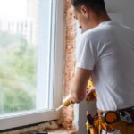A man wearing a tool belt is pointing a caulk gun at the bottom of a window frame. The insulation is exposed.