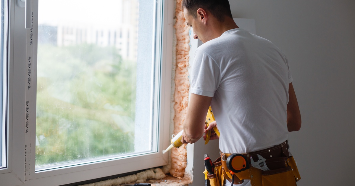 A man wearing a tool belt is pointing a caulk gun at the bottom of a window frame. The insulation is exposed.