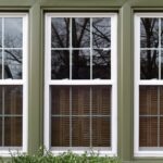 Three double-hung windows with green trim on a beige home, reflecting trees and the sky, with bushes below.