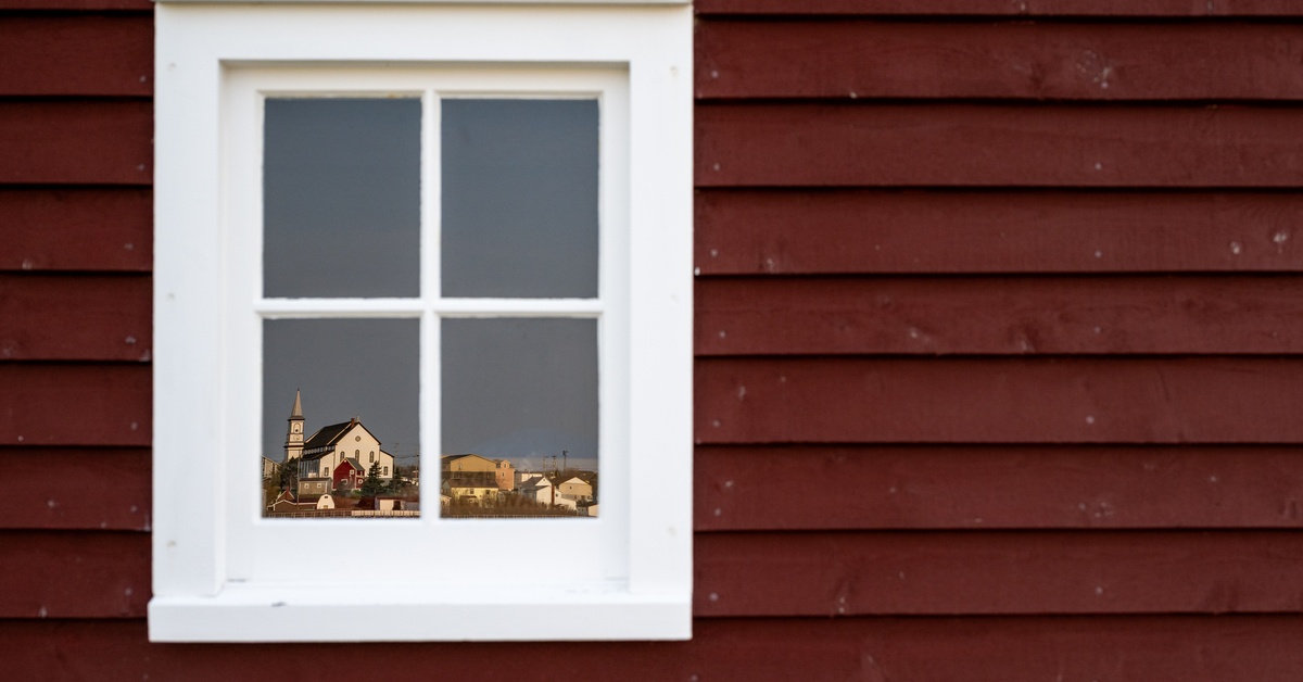 A 4-paned window with a white frame on a home with dark red wooden siding. The window shows a reflection of a historical town.