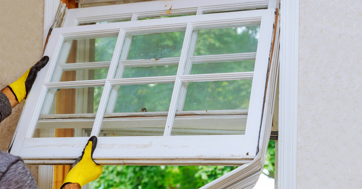 A person wearing yellow work gloves is tearing a multi-pane sash window out of its wooden frame with green foliage outside.