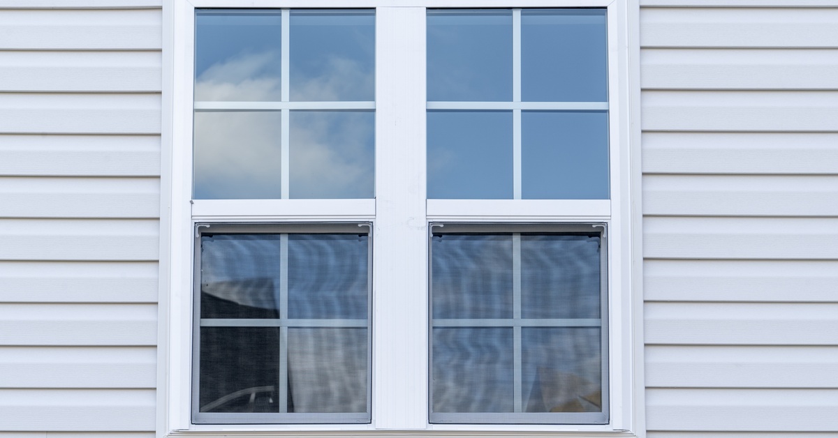 A white vinyl-framed double-hung window on a house with white siding. The sky is reflected in the window.