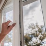 A hand grips the handle of a white vinyl window, partially opening it. Trees and a cloudy sky are blurred outside.
