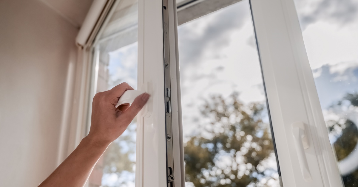 A hand grips the handle of a white vinyl window, partially opening it. Trees and a cloudy sky are blurred outside.