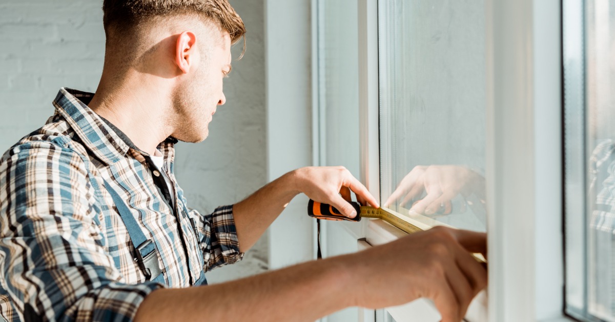 A person squints in the sun as they use a tape measure to take the dimensions of a white window frame.