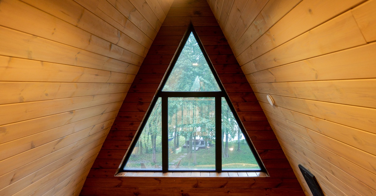 The interior of an A-frame cabin with a triangular window overlooking lush green trees and another building in the distance.