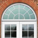 A red brick wall with French doors and a rounded accent window, framed by lanterns. A small plaque is mounted to the wall.