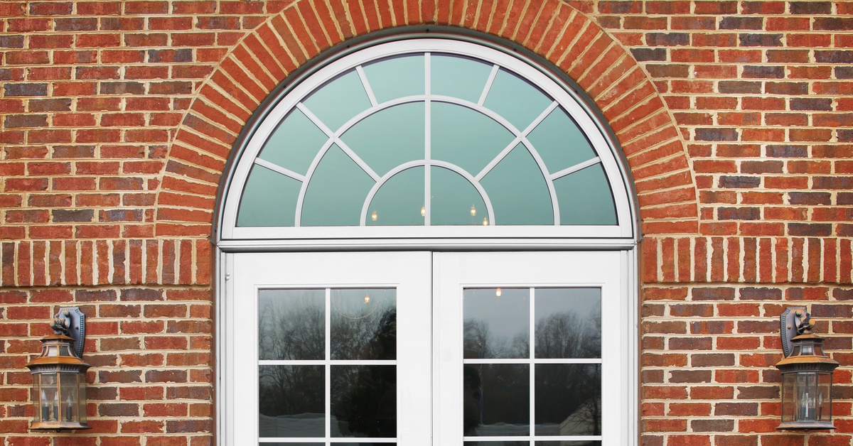 A red brick wall with French doors and a rounded accent window, framed by lanterns. A small plaque is mounted to the wall.