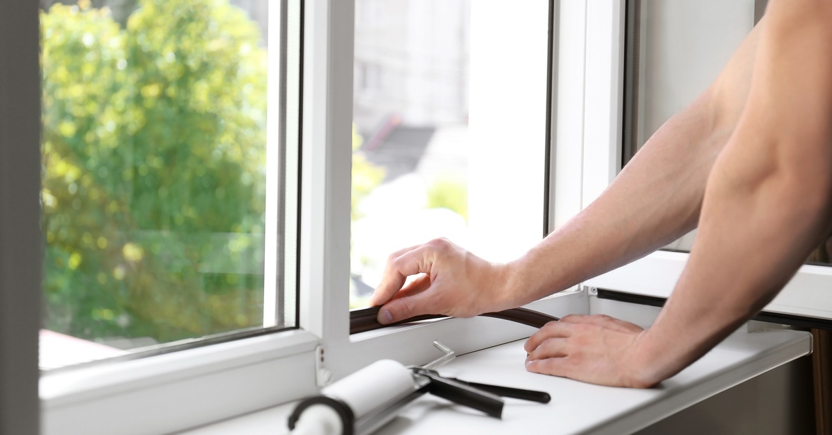 Person applies weatherstripping to a window to improve energy efficiency. A caulking gun sits on the windowsill.