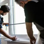 Two installers measure a window frame inside a home while preparing the opening for a new window installation.