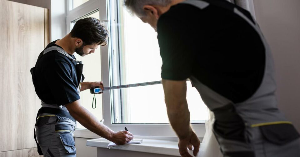 Two installers measure a window frame inside a home while preparing the opening for a new window installation.