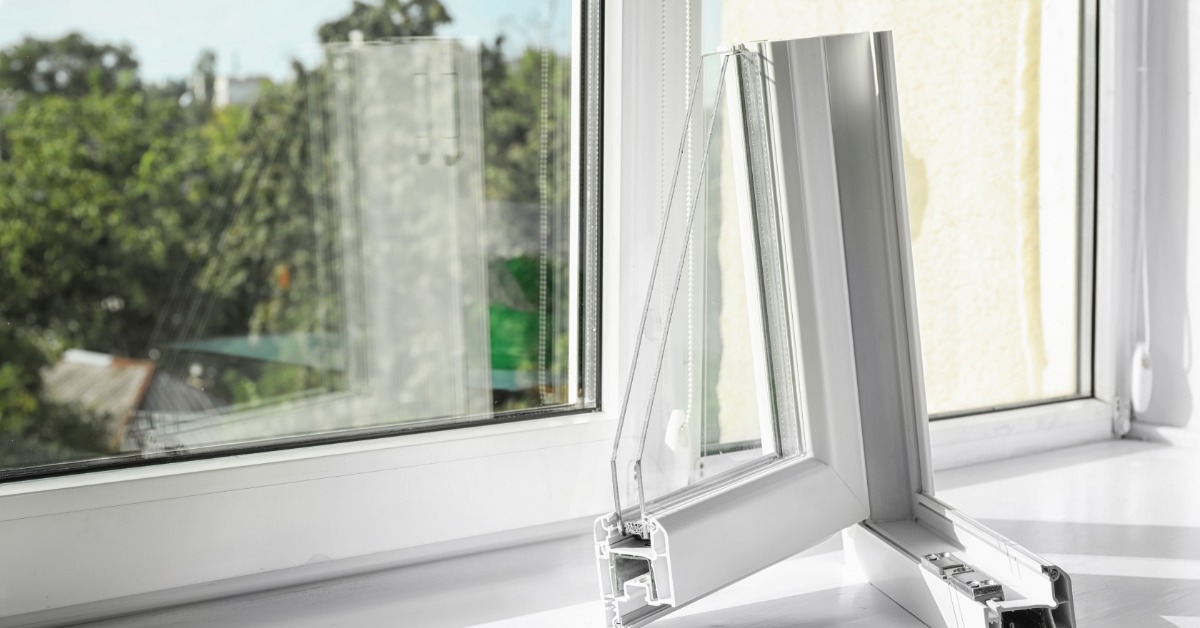 A cutaway sample of a vinyl window frame sits on a windowsill, showing internal chambers with a window and greenery outside.