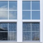 A white double-hung window with grid panes is set into light-colored vinyl siding, reflecting blue sky and clouds outside.