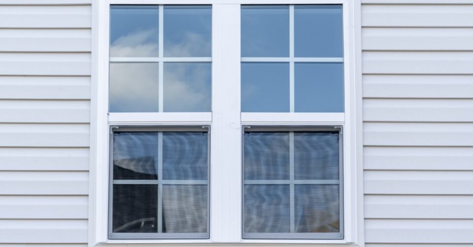 A white double-hung window with grid panes is set into light-colored vinyl siding, reflecting blue sky and clouds outside.
