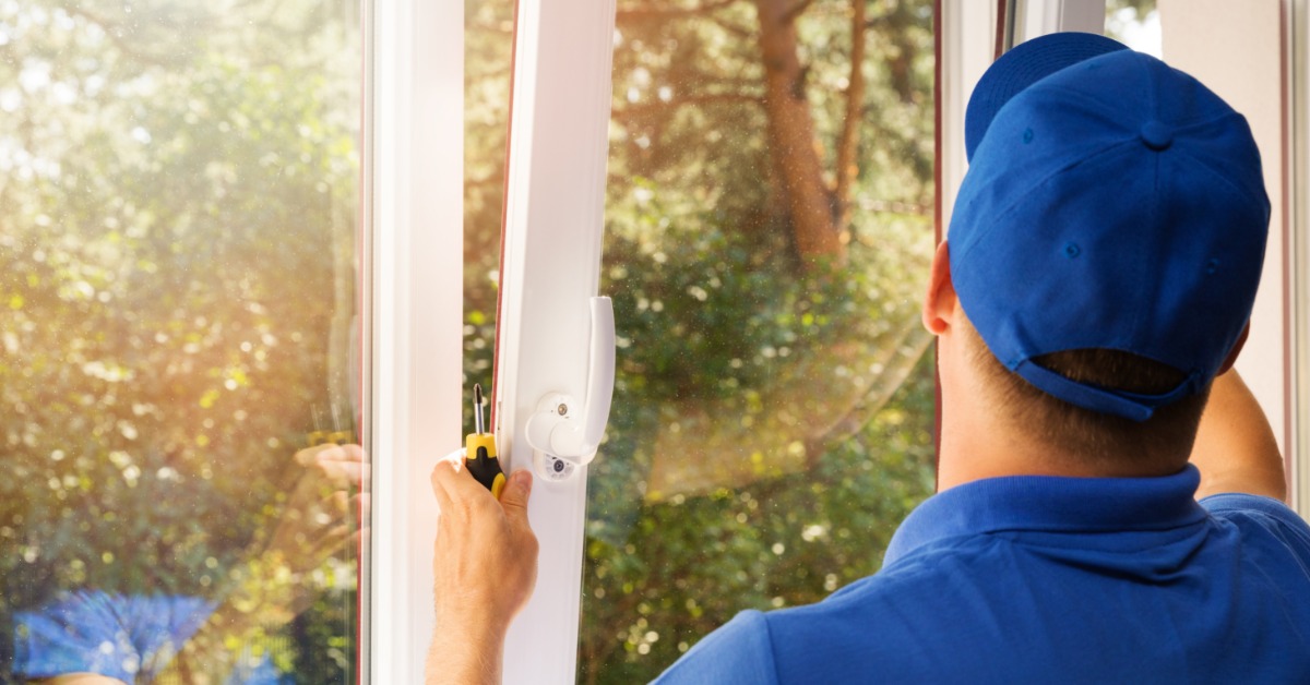 A man in a blue hat and uniform installing a white window into a home looking out at a dense treeline.