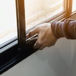 a person wearing a gray sweater installing an aluminum screen window frame alongside a white wall indoors.