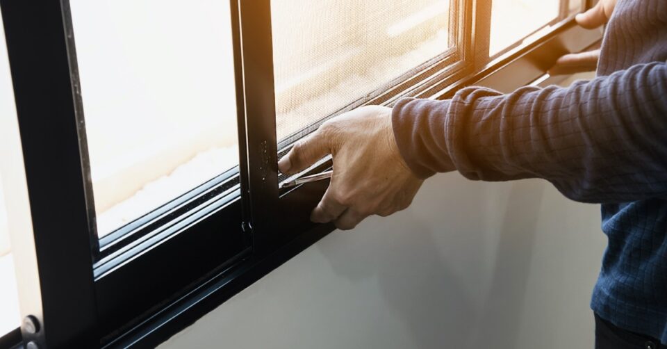 a person wearing a gray sweater installing an aluminum screen window frame alongside a white wall indoors.