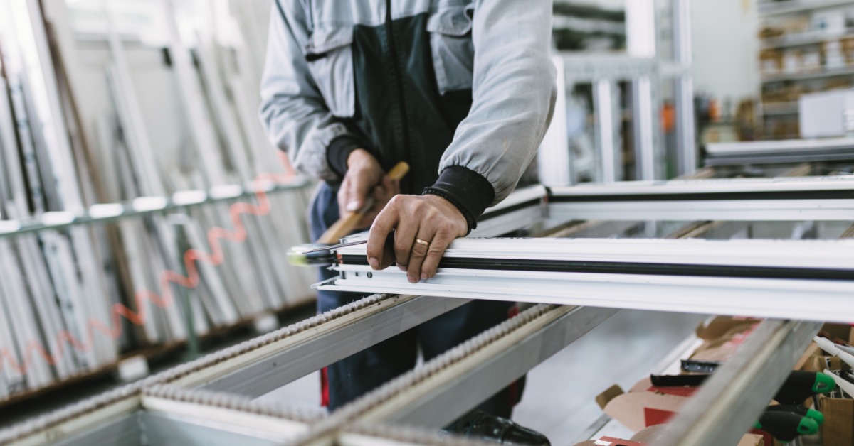 a worker assembling a white aluminum window or door frame with tools and equipment inside a manufacturing facility.