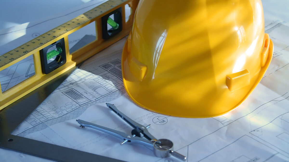 a set of home plans with a hard hat, compass, and level on a wooden work surface beside a ruler.