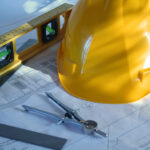 a set of home plans with a hard hat, compass, and level on a wooden work surface beside a ruler.