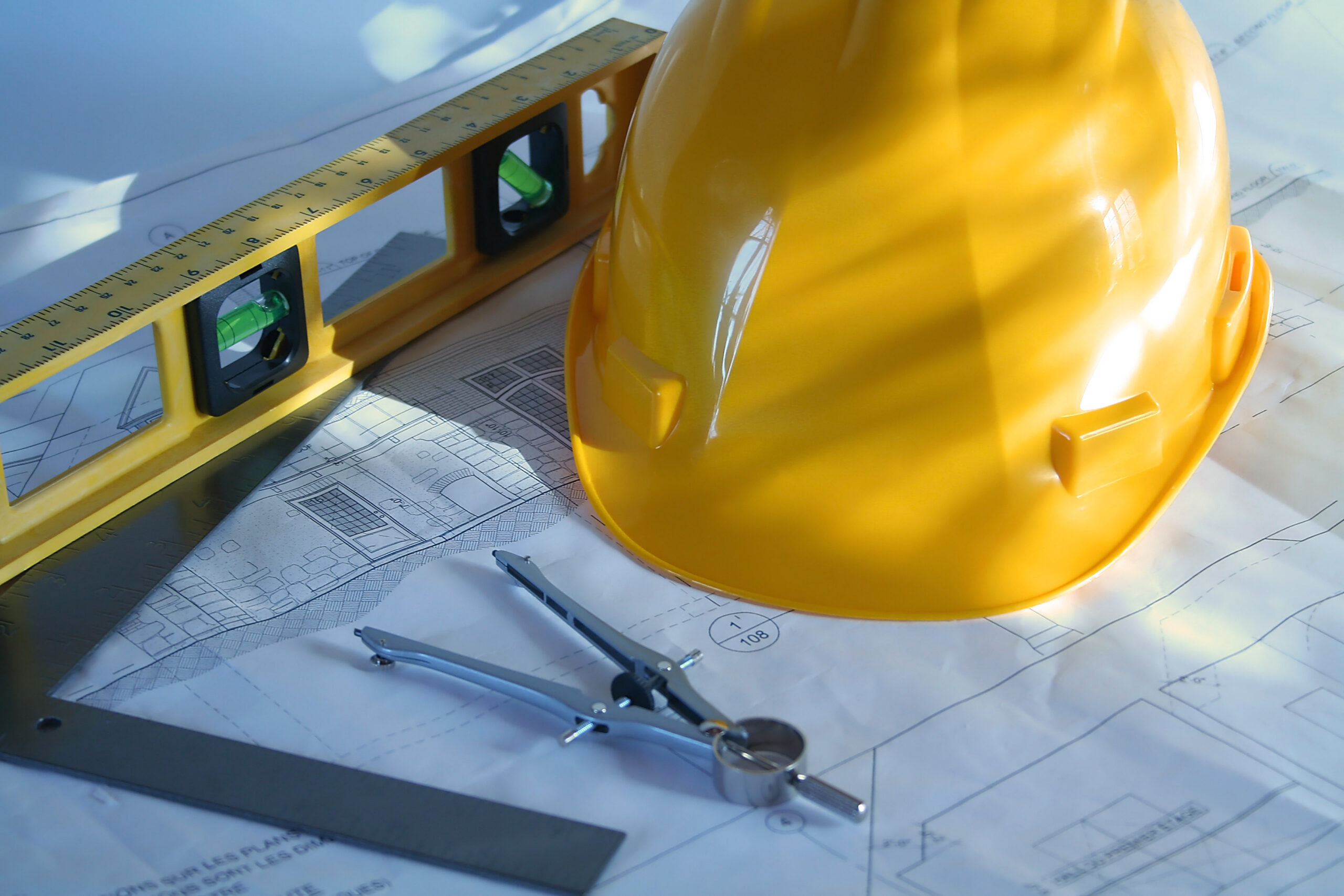 a set of home plans with a hard hat, compass, and level on a wooden work surface beside a ruler.