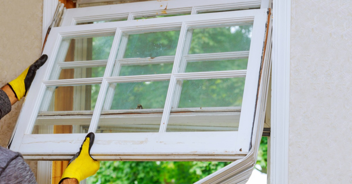 Yellow-gloved hands holding an older white window sash inside a wall opening with green trees outside.