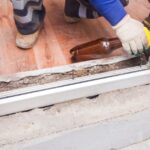 A gloved hand using a yellow caulk gun at the base of a white window with tools and flooring nearby.