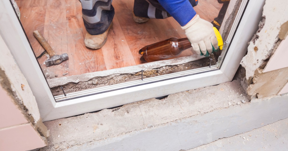A gloved hand using a yellow caulk gun at the base of a white window with tools and flooring nearby.