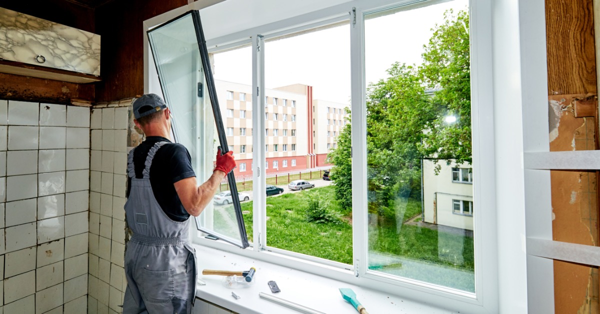 A person installing a white window inside a room with tiled walls, tools on the sill, and buildings outside.