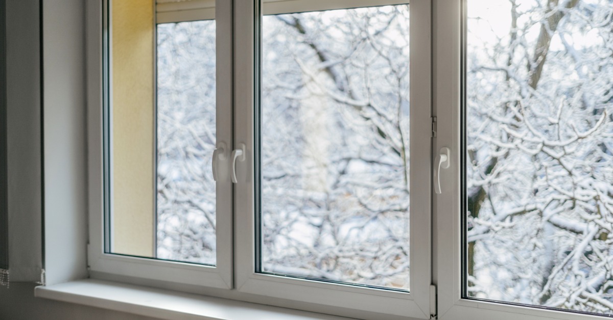 A white-framed window with multiple glass panes overlooking snow-covered trees from inside a room with a white sill.